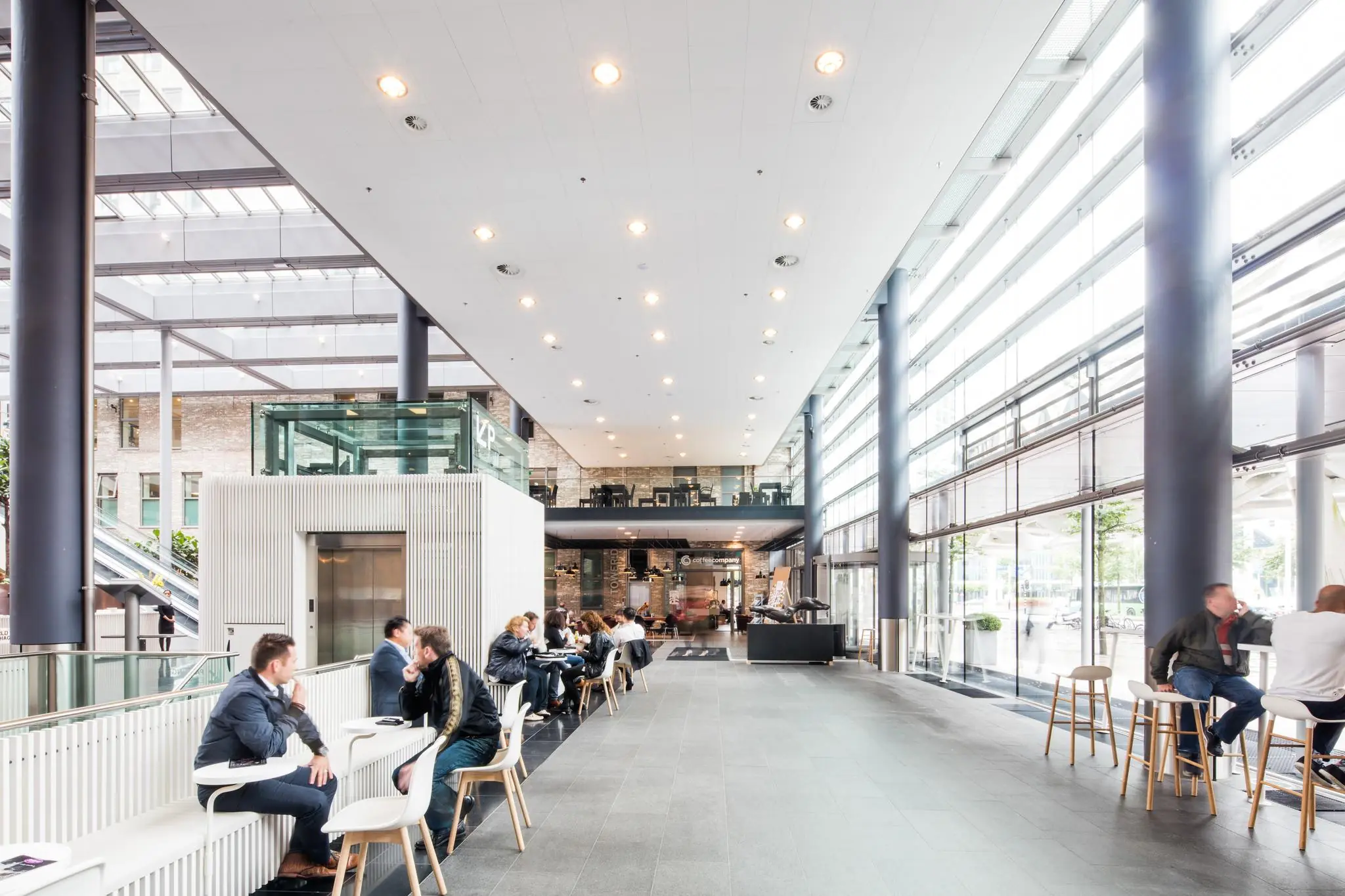 Moderne, lichte kantoorlobby aan de Prinses Beatrixlaan met mensen die aan tafeltjes zitten en een koffiebar op de achtergrond.